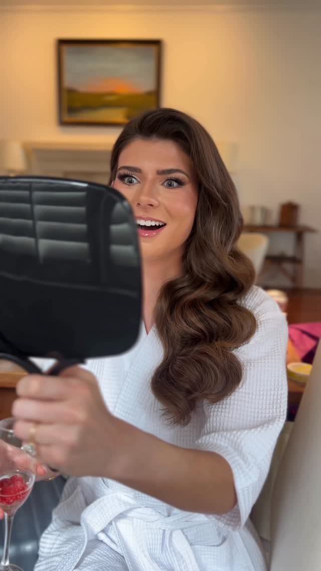 Woman with a deep side part and wavy brown hair looking into a mirror.