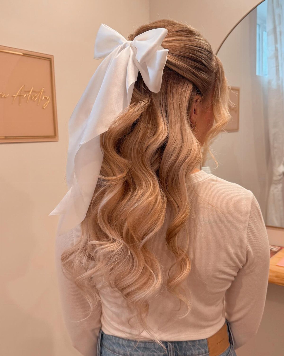 Blonde wavy hair styled half-up with a large white fabric bow accessory.
