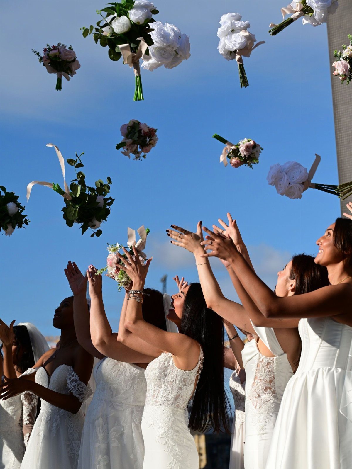 Women in white wedding dresses reaching for floating floral bouquets against a blue sky