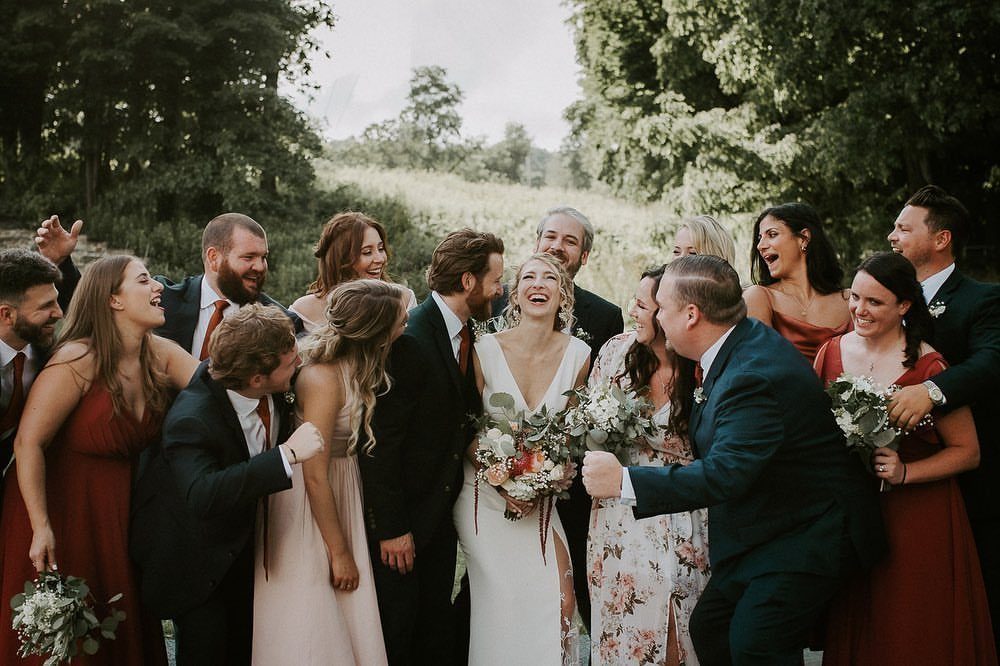 Bridal party laughing and cheering around a bride and groom in a field