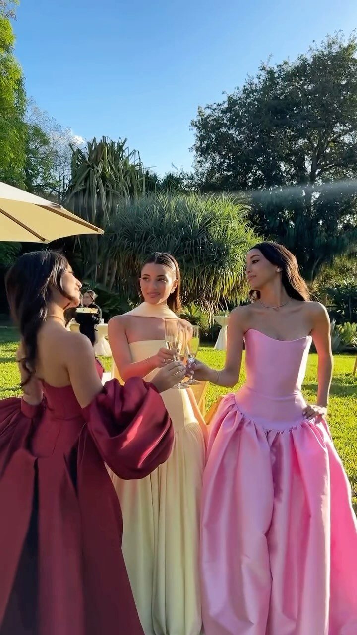 Three women in colorful formal gowns holding champagne glasses in a sunny garden.