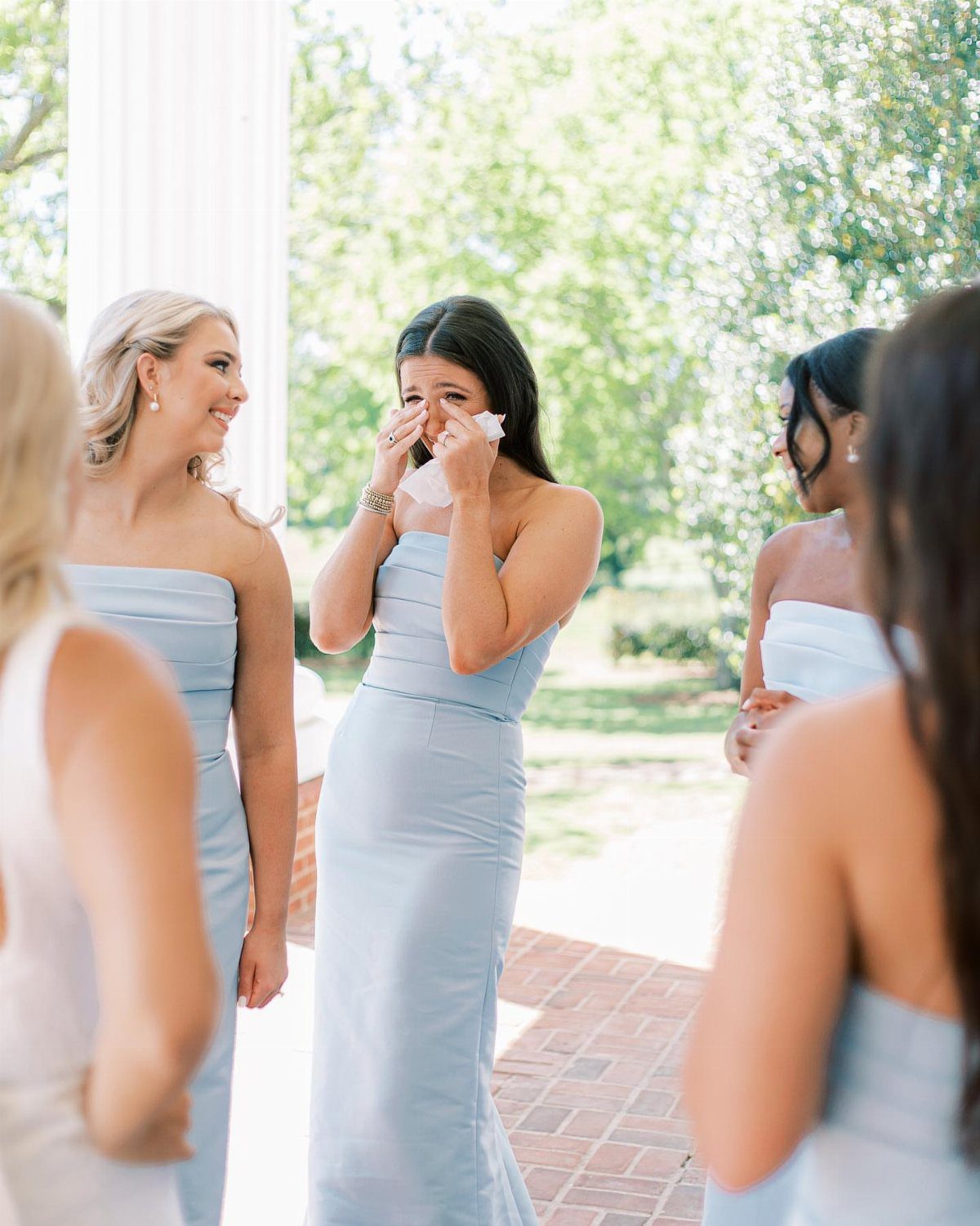 Bridesmaids in light blue dresses standing together, one woman dabbing her eyes.