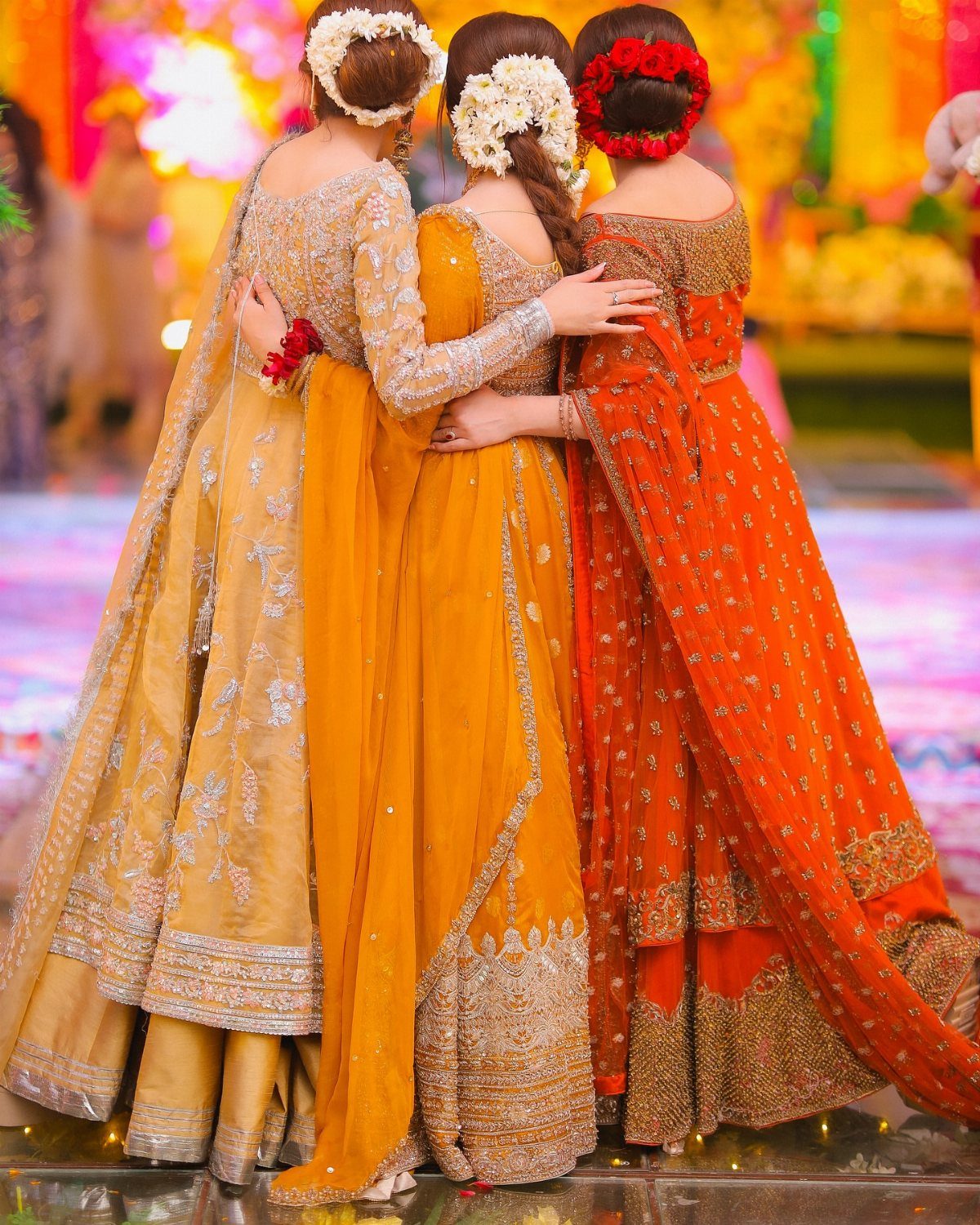 Three women in colorful traditional South Asian dresses embracing in a group hug.