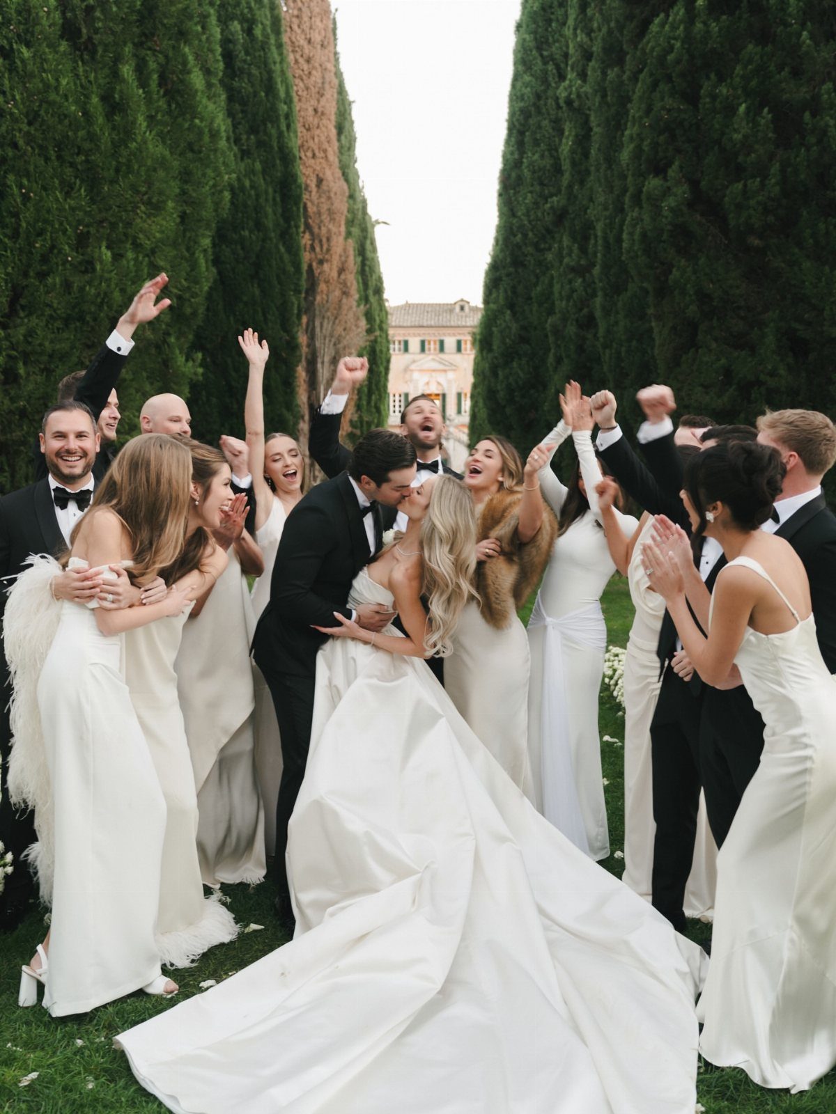 Bride and groom kissing while their bridal party cheers around them outdoors.
