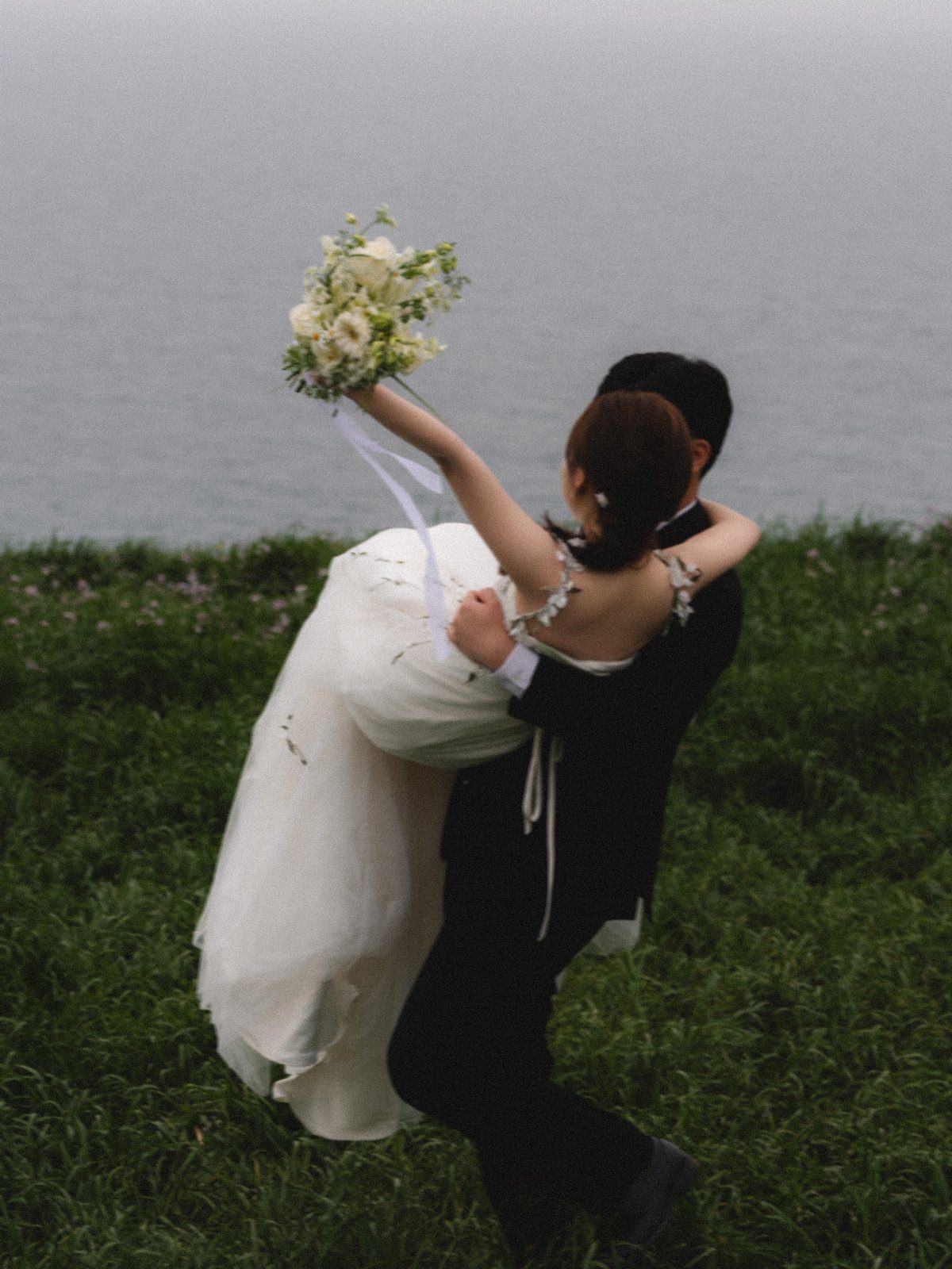 Groom carrying bride sideways in his arms on a grassy cliff by the sea