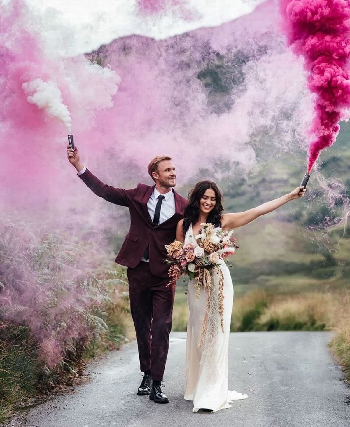 Bride and groom holding smoke bombs emitting pink and white clouds on a road