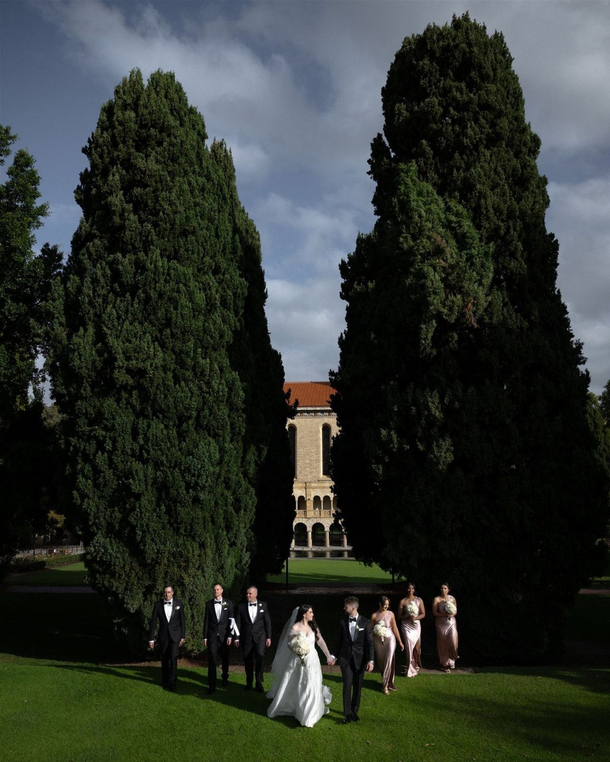 Bridal party walking across a green lawn between two large cypress trees.