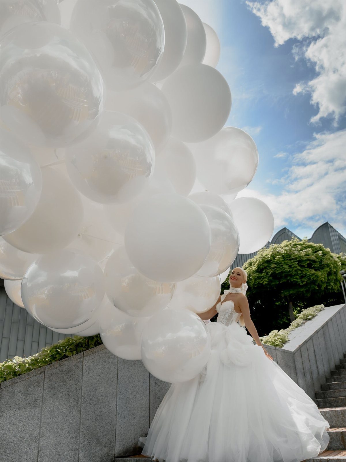 Bride in a white wedding dress holding a large cluster of white balloons outdoors.