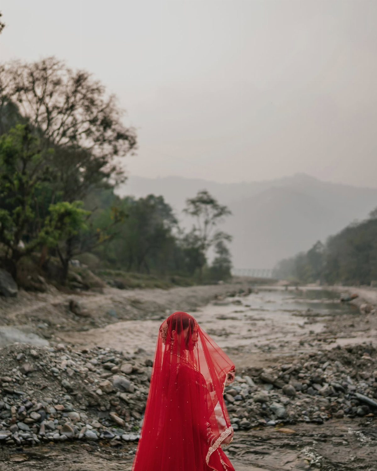 Bride in red veil standing in a rocky riverbed with misty mountains behind.