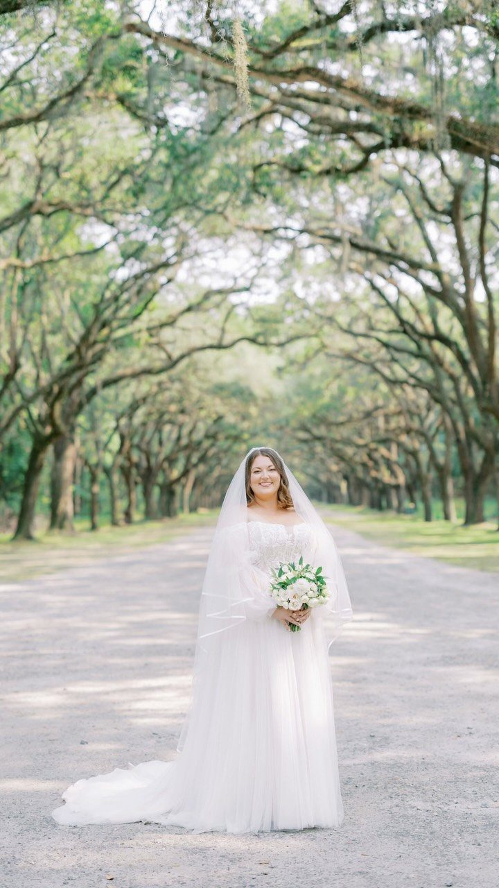 Bride in a white dress holding a bouquet on a tree-lined path.
