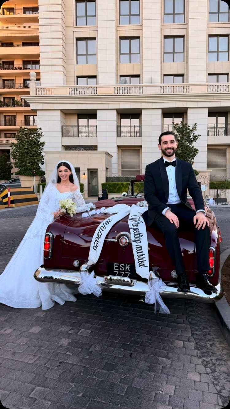 Bride and groom posing with a maroon vintage car decorated with white ribbons.