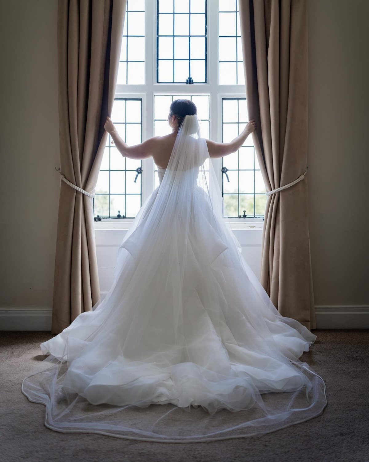 Bride in white gown opening tan curtains to look through a large window