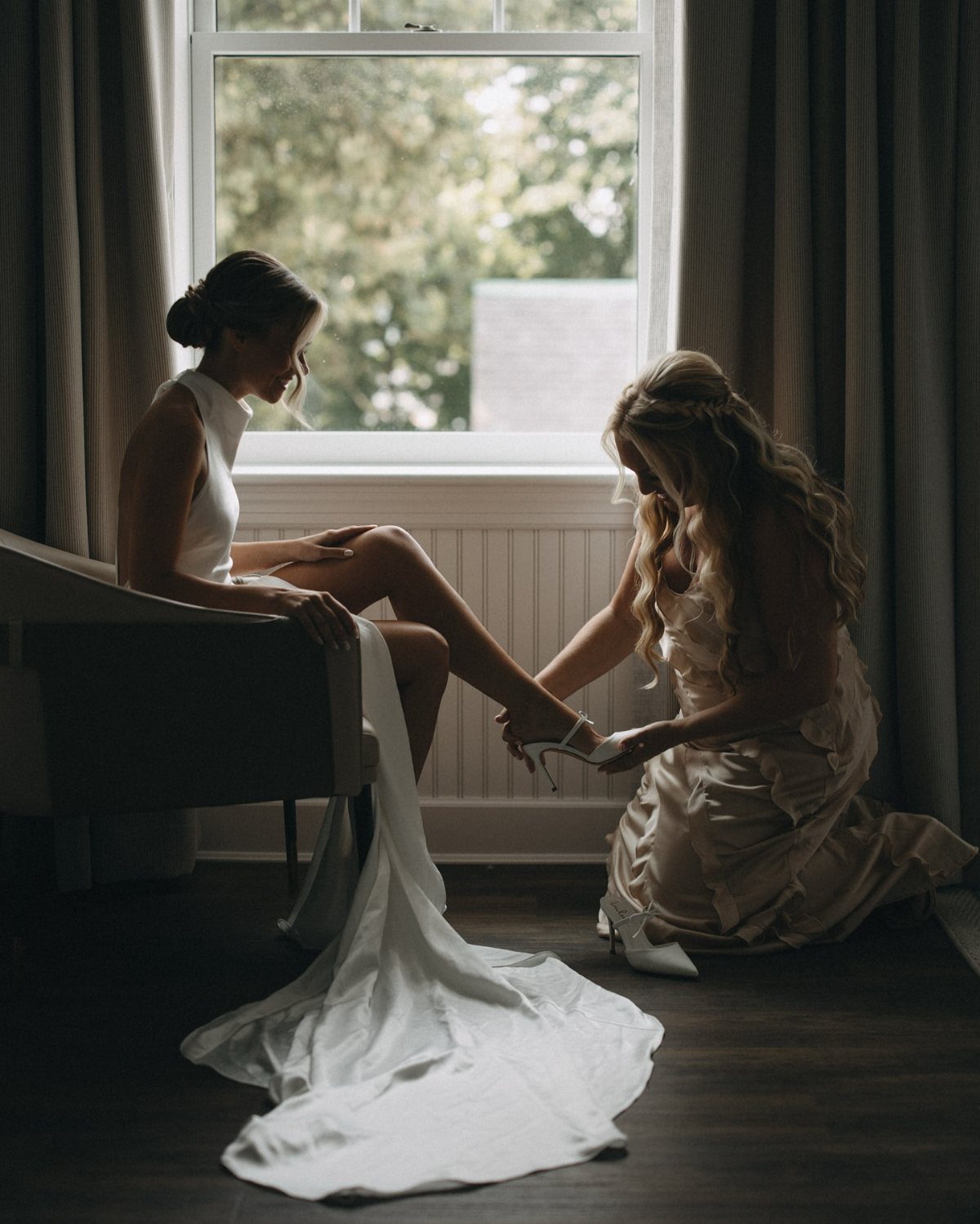 Bride sitting by a window while a bridesmaid helps her put on shoes.