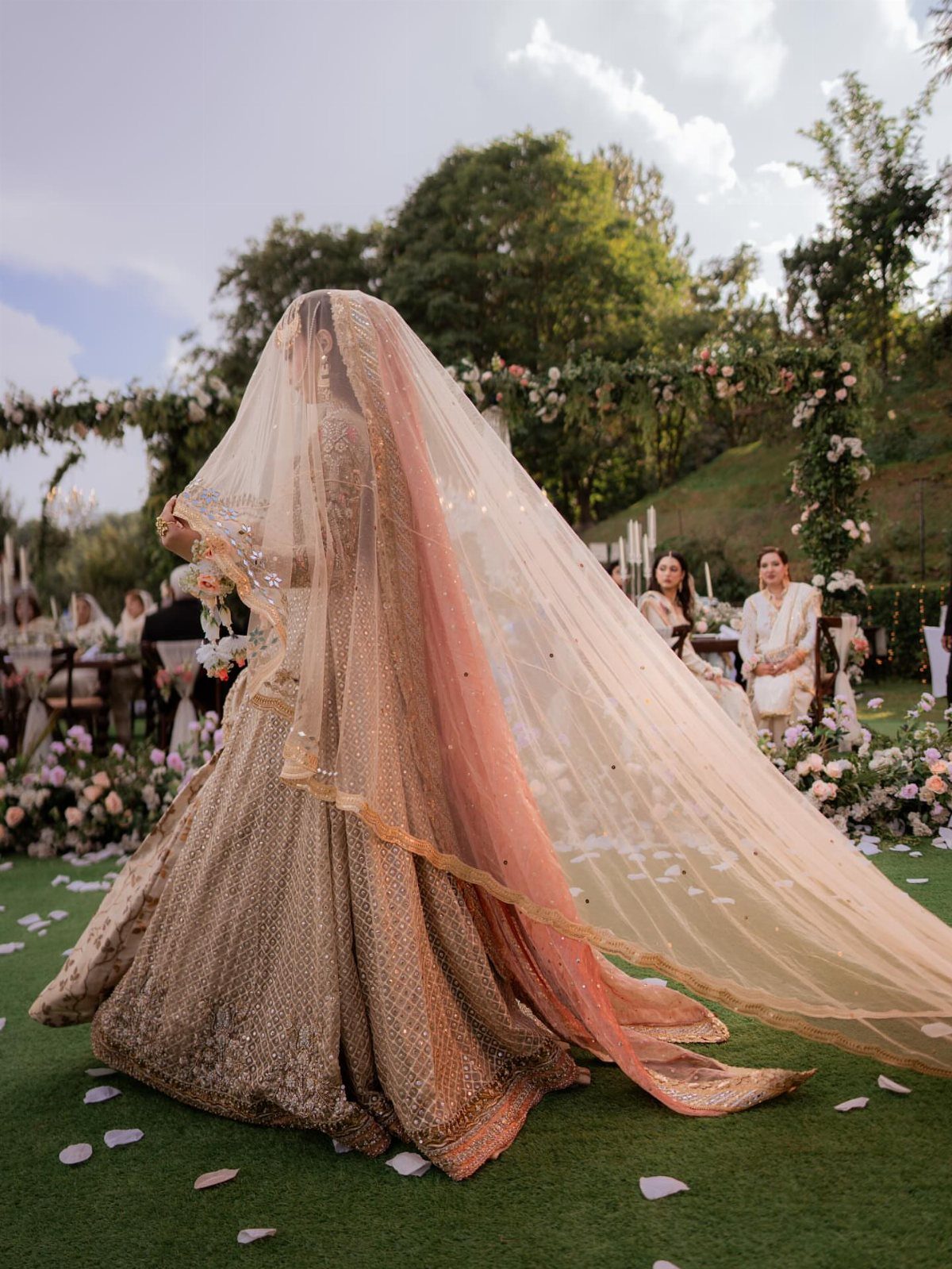Bride in gold lehenga with a peach dupatta billowing in the wind outdoors