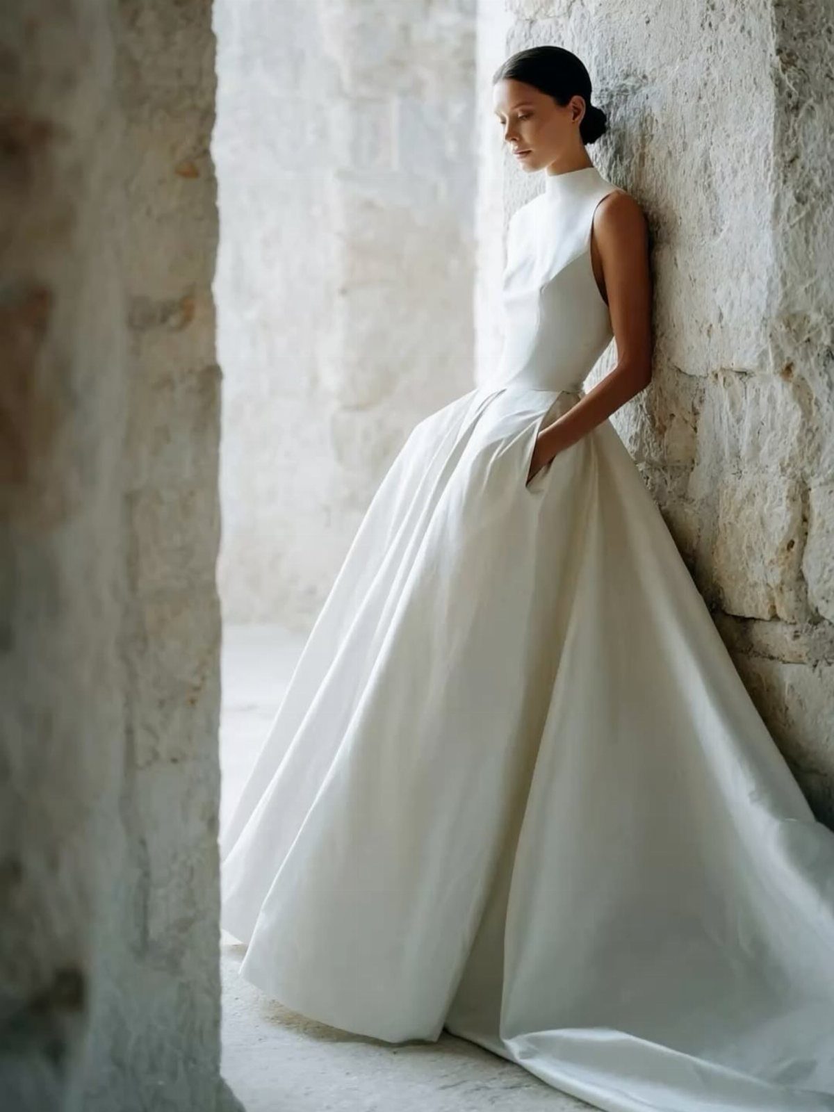 Bride in a white gown leaning against a stone wall with hand in pocket