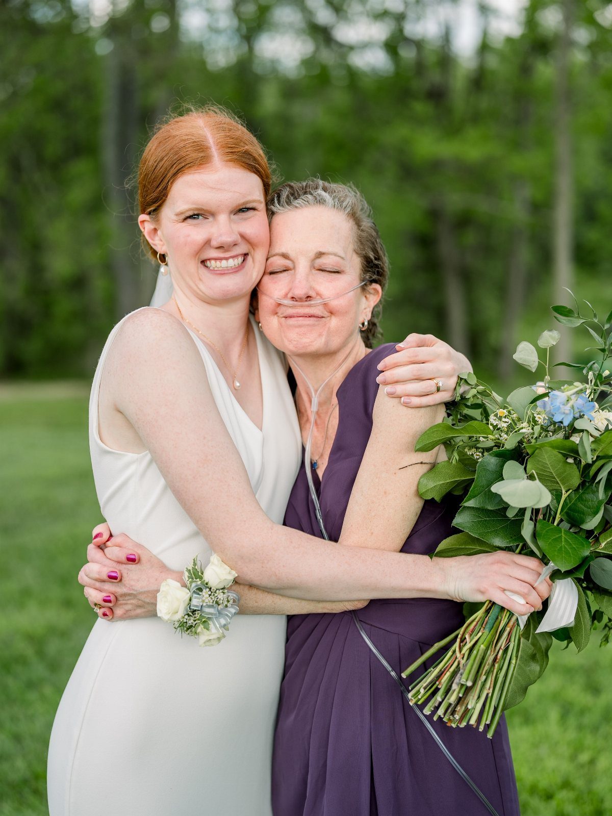 Bride in white dress hugging an older woman in a purple dress outdoors.