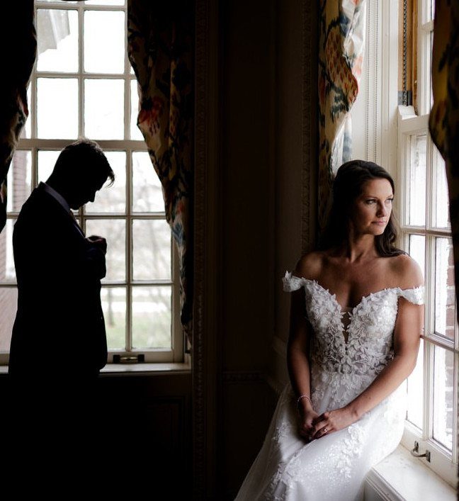 Bride in white lace dress sitting by a window with a silhouetted groom.
