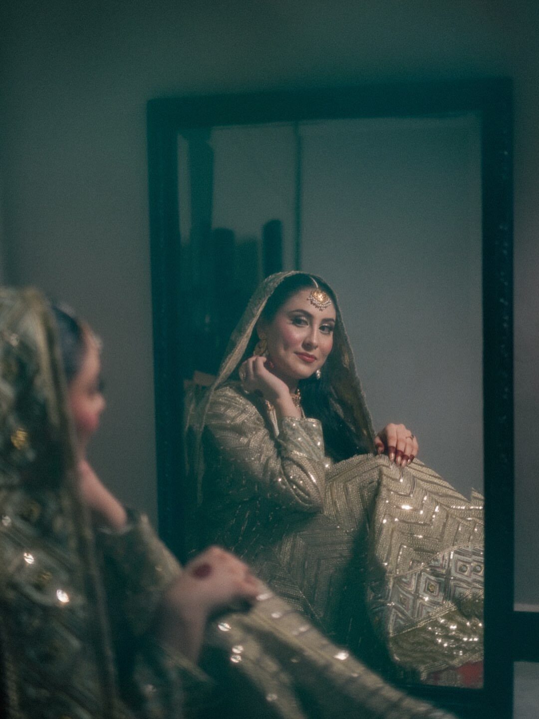 Bride in gold lehenga smiling at her reflection in a dark-framed mirror