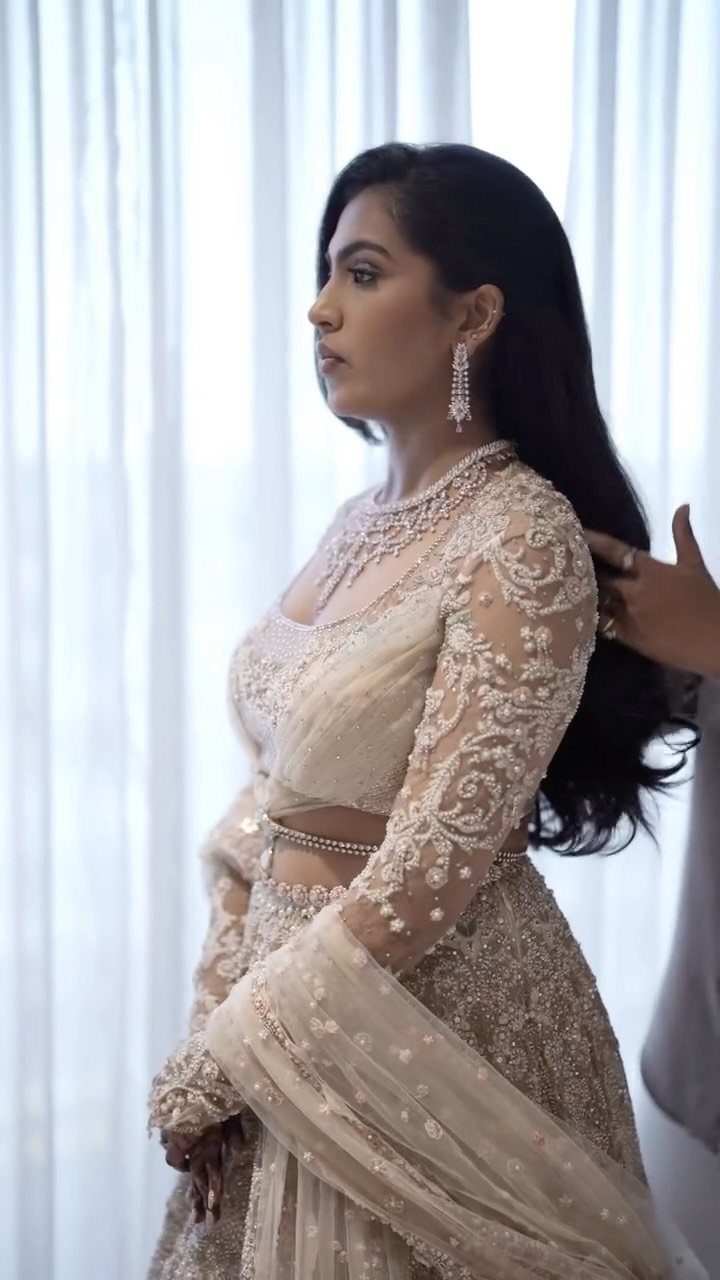 Woman in an embroidered beige lehenga posing in profile against white curtains.