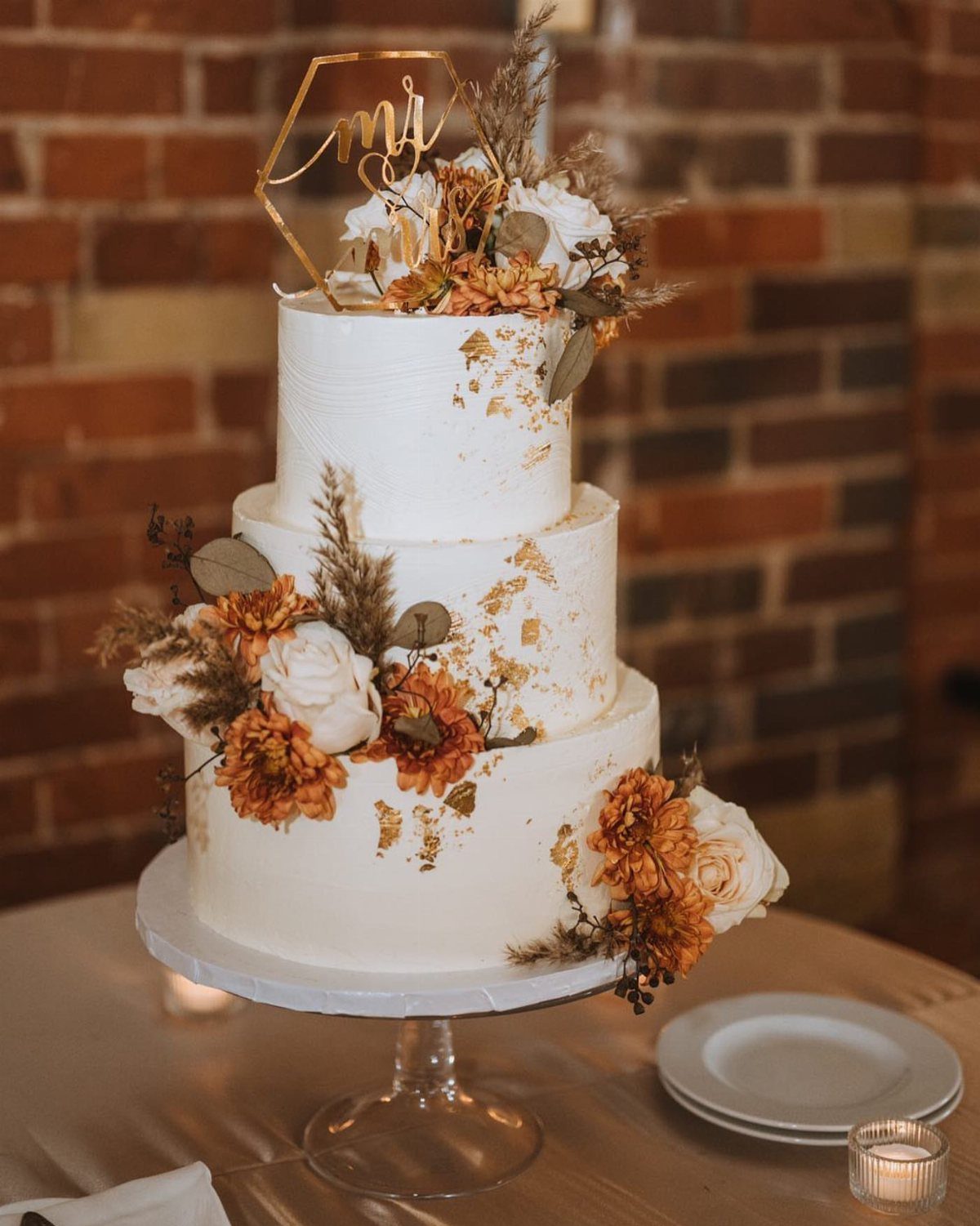 Three-tier white buttercream wedding cake with gold leaf and autumn floral decorations.