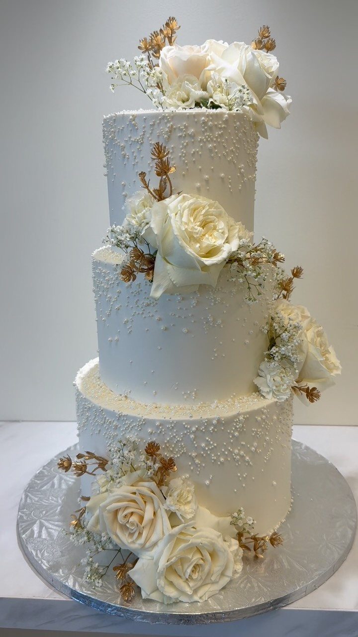 Three-tier white buttercream wedding cake decorated with white roses and edible pearls.