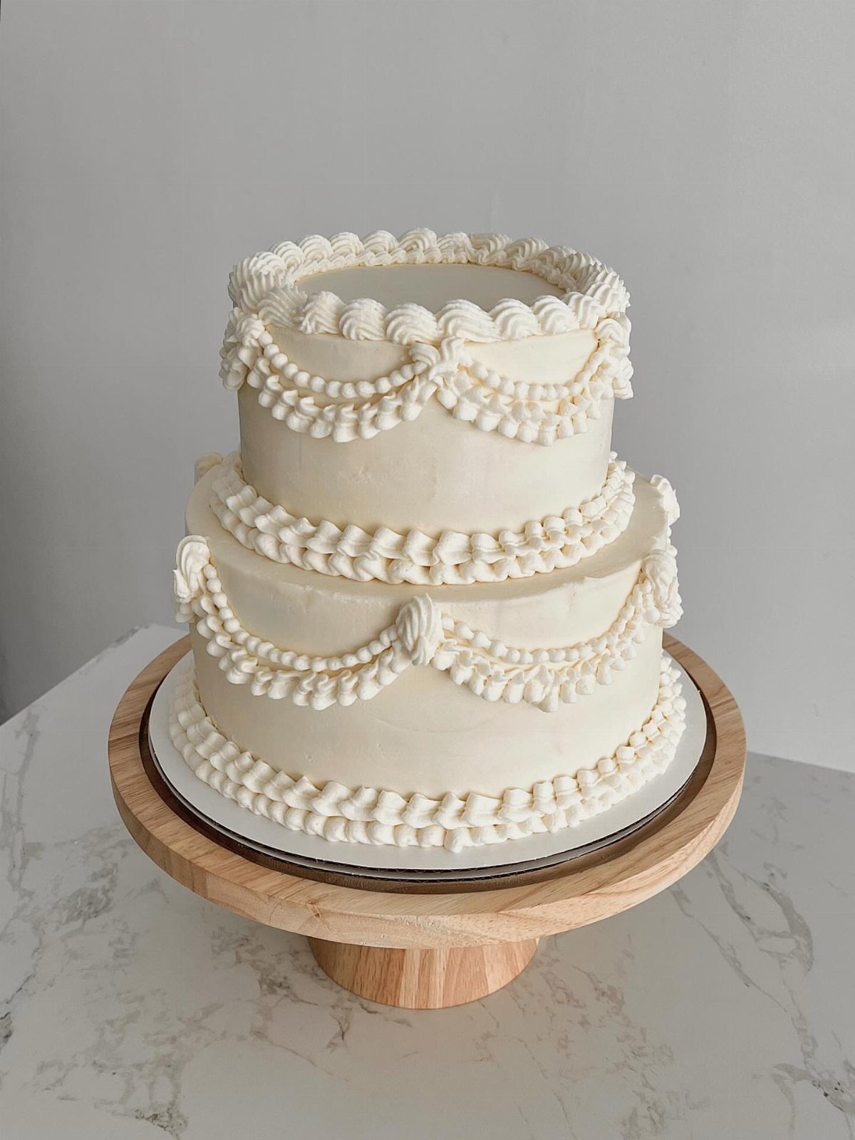 Two-tier white buttercream wedding cake with decorative piped swags on a wooden stand.