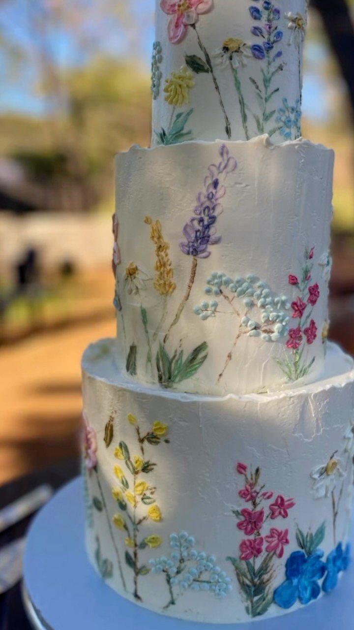 Three-tiered white buttercream cake decorated with colorful piped wildflowers on a white stand.