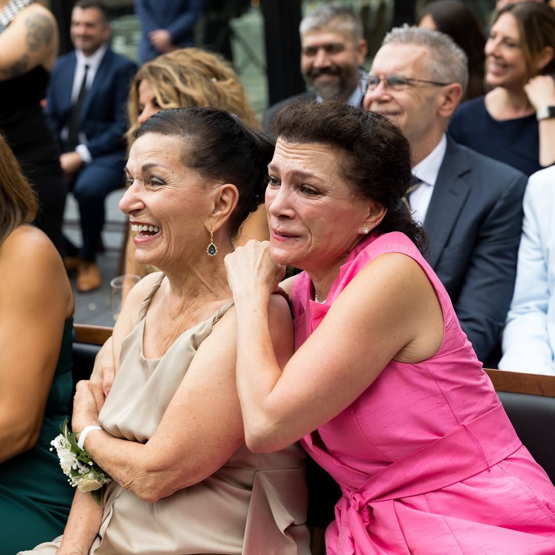 Two women reacting emotionally, one laughing and one crying, at a wedding ceremony.