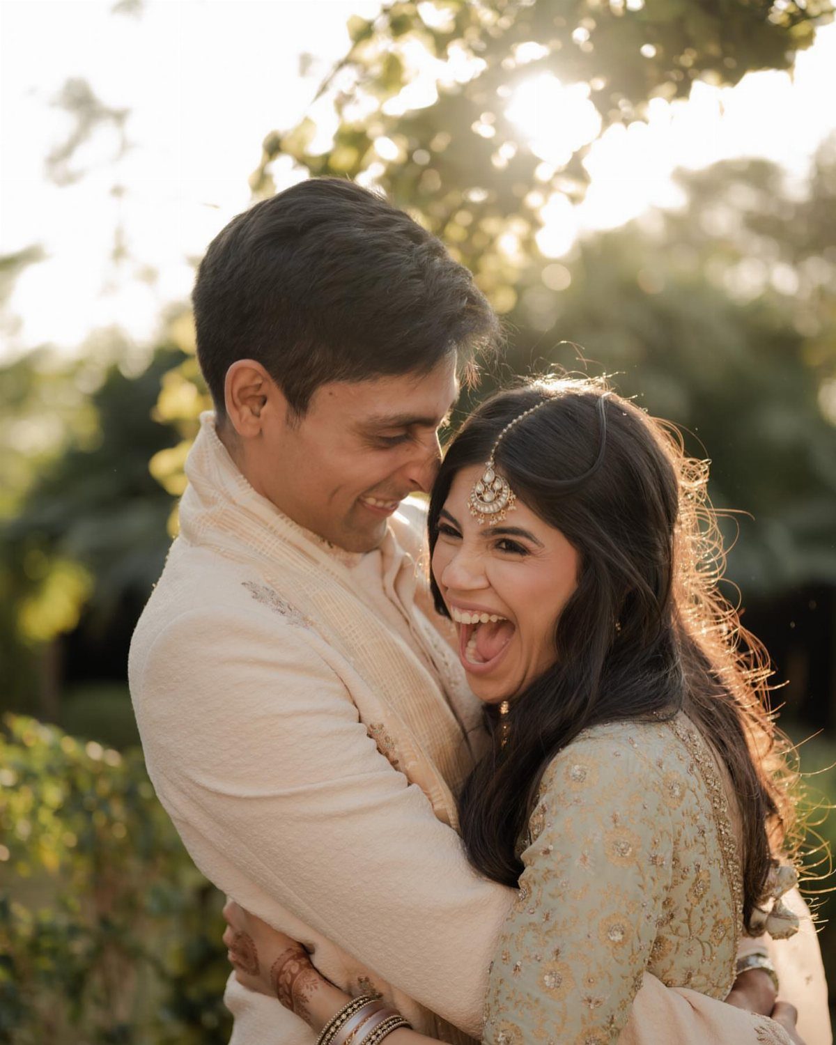 Couple in Indian wedding attire embracing and laughing joyfully in a sunlit garden