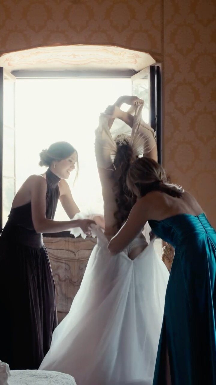 Bridesmaids helping a bride put on her white wedding dress in a room.