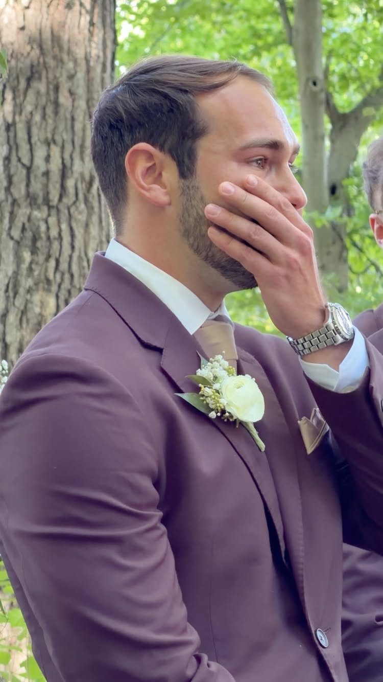 Groom in a mauve suit wiping a tear from his eye outdoors.