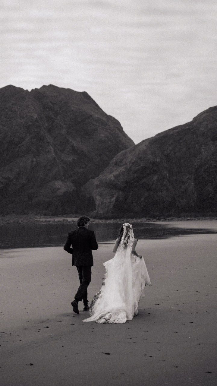 Bride and groom walking away on a beach toward large mountains in monochrome.