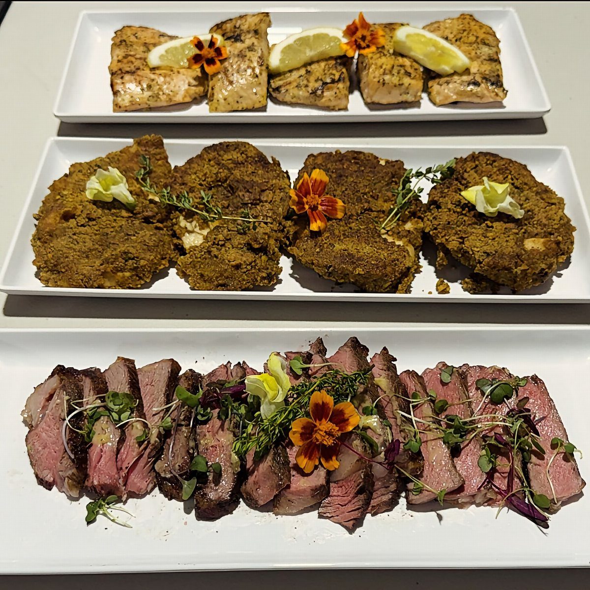 Three platters of grilled salmon, breaded cutlets, and sliced steak garnished with flowers.