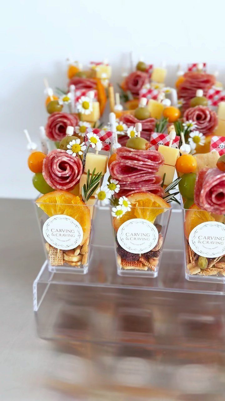 Appetizer cups with rolled salami, cheese, olives, and crackers on a clear riser.