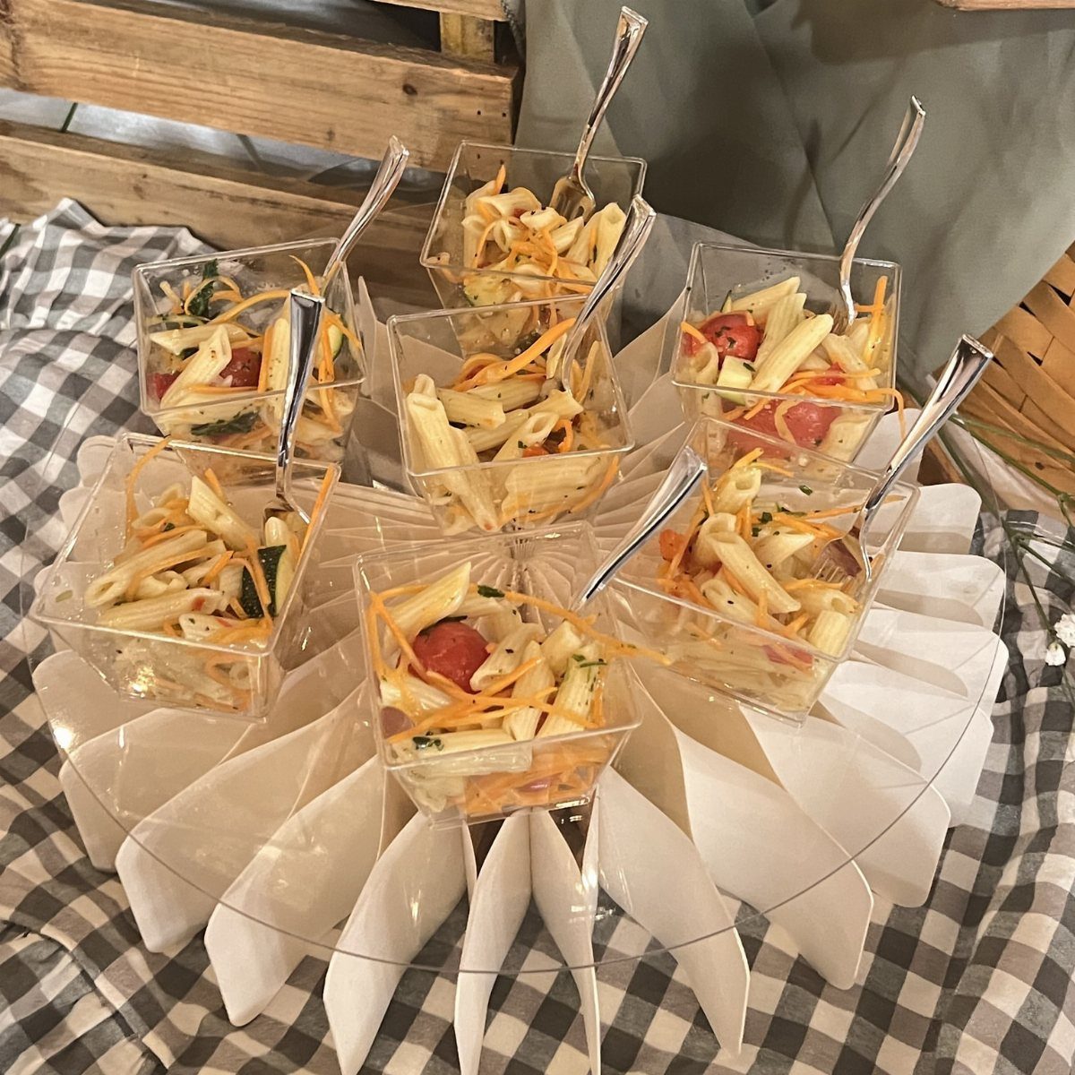 Penne pasta salad in small clear plastic cups with silver forks on a stand.