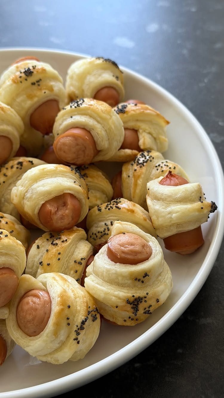 Mini sausages wrapped in golden puff pastry with poppy seeds on a white plate.