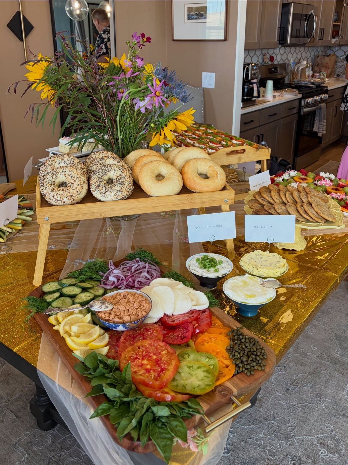 Food spread with bagels, fresh vegetable platter, and wildflowers on gold tablecloth