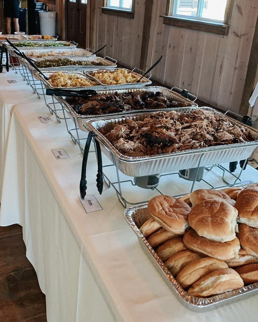 Buffet table with pulled pork, grilled meats, sides, and dinner rolls in trays.