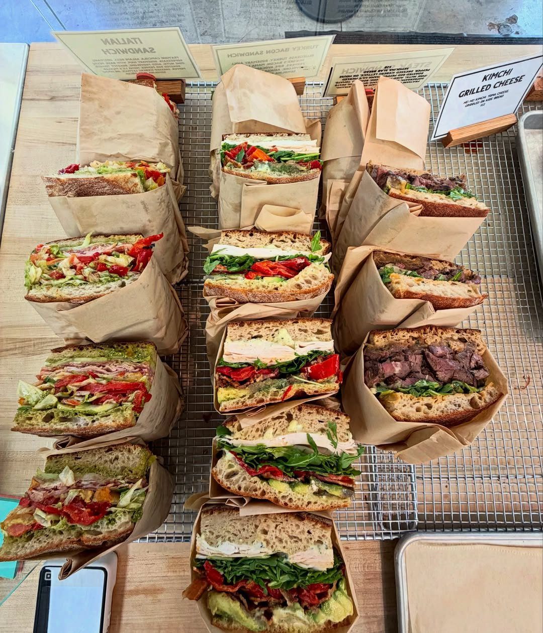 Assorted gourmet sandwiches wrapped in brown paper on a metal wire rack.