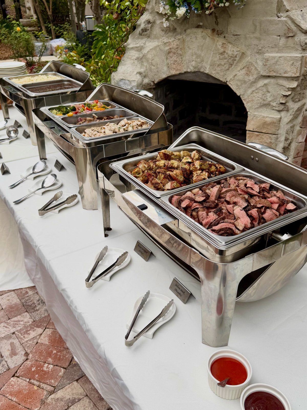 Buffet line with sliced meat and roasted potatoes in silver chafing dishes outdoors.