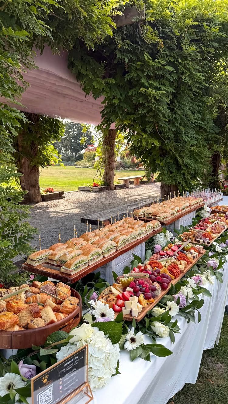 Buffet table with sandwiches, fruit, and pastries decorated with white flowers outdoors.