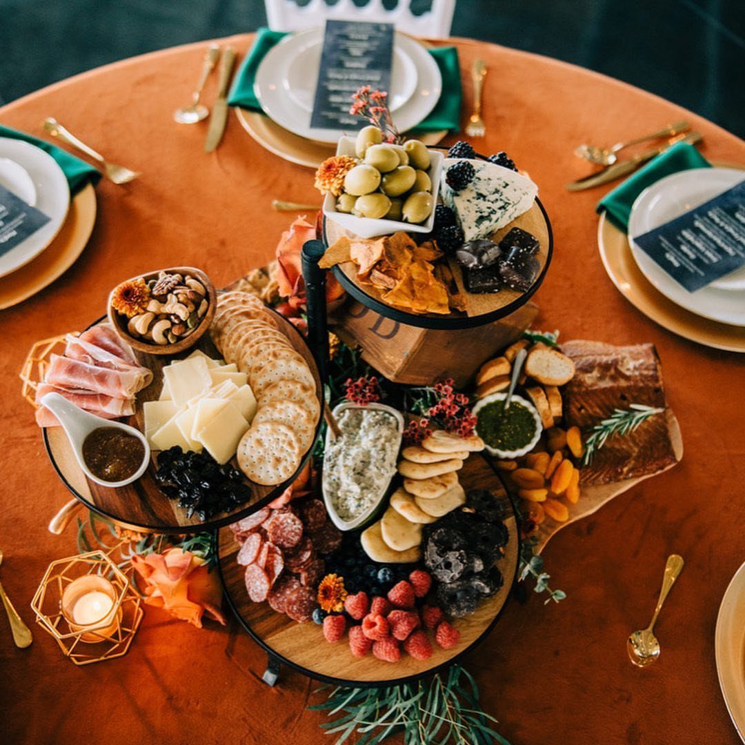 Tiered grazing table with cheeses, meats, and fruits on an orange tablecloth.