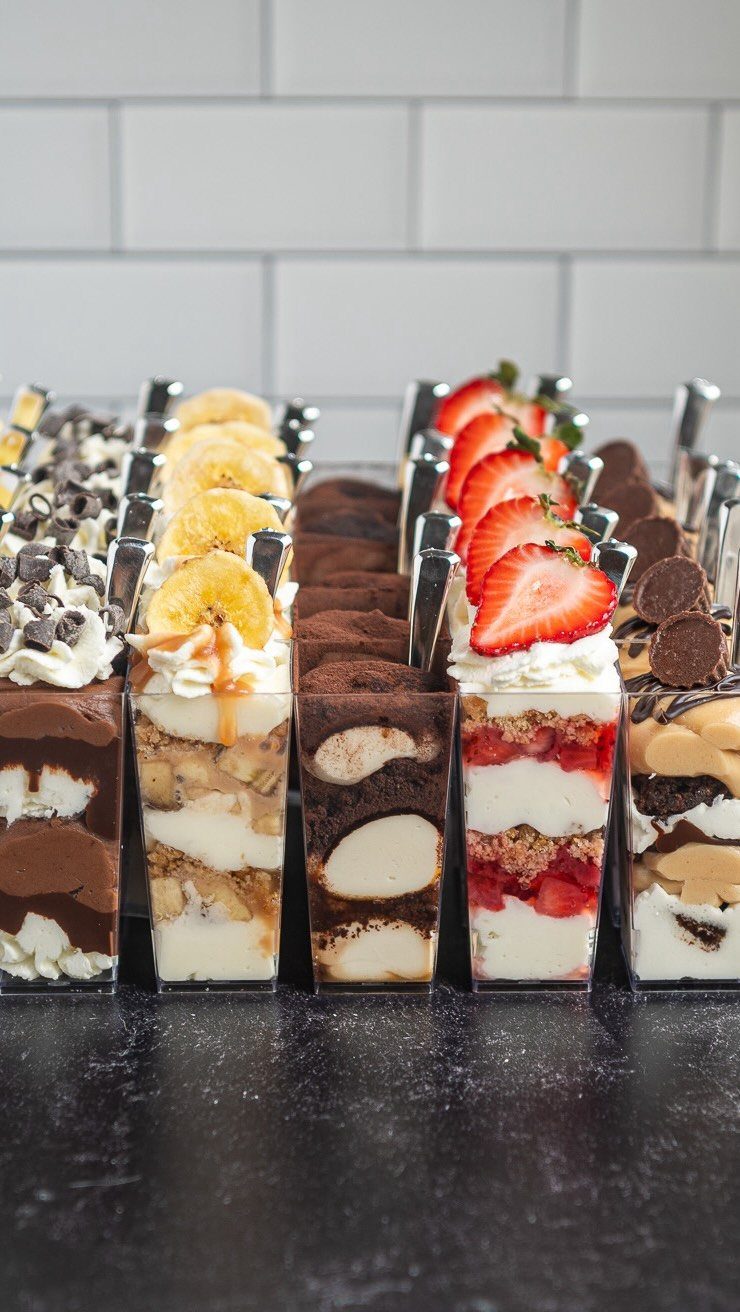 Layered cheesecake dessert cups with strawberry, banana, and chocolate toppings on a dark surface.