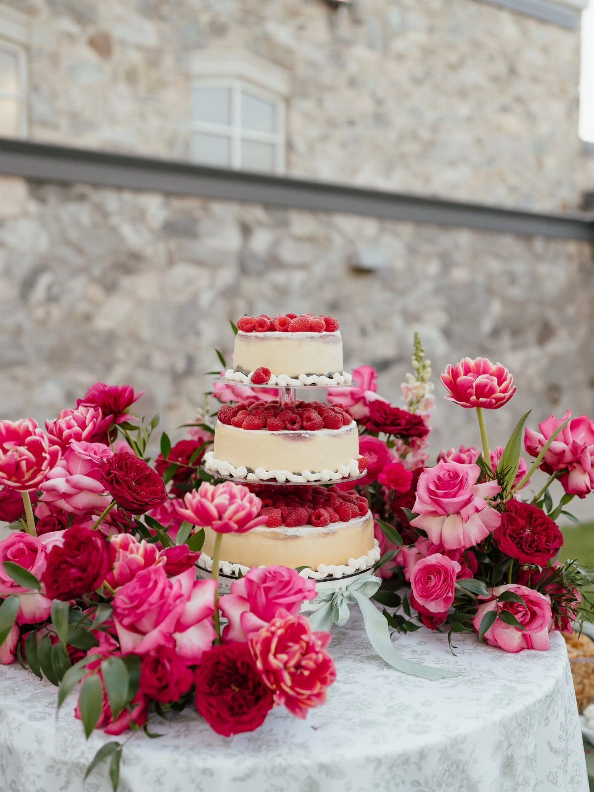 Three-tiered raspberry cake surrounded by pink and red roses on a white table.