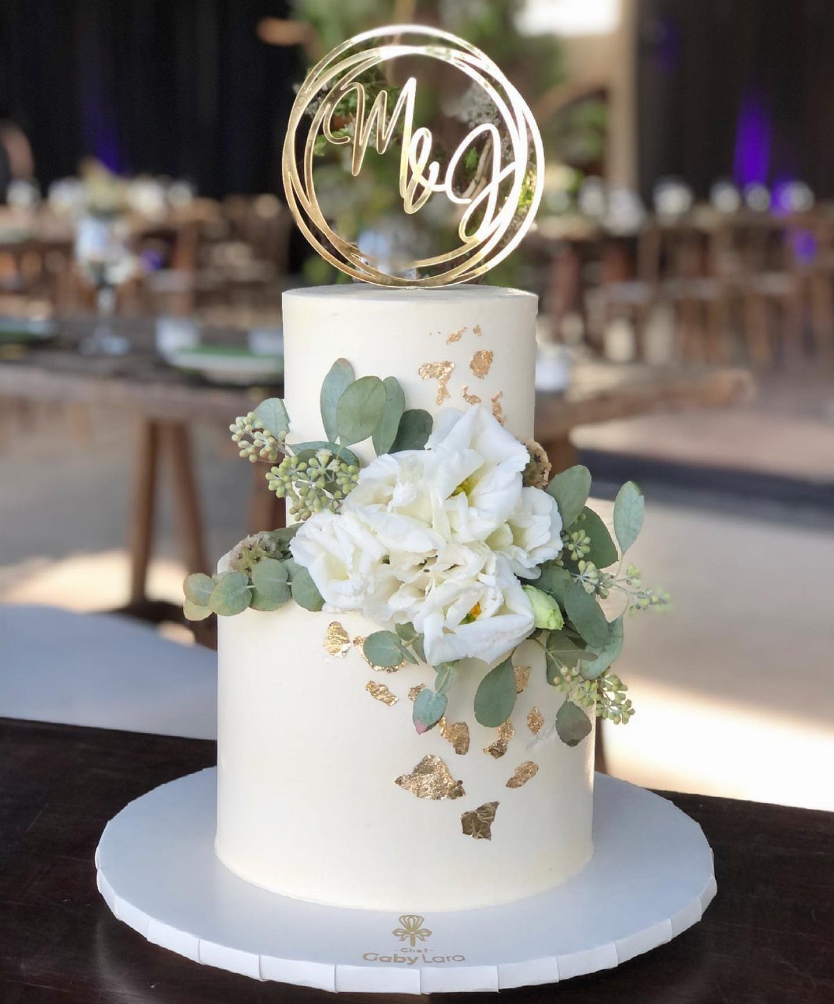 Two-tier white wedding cake decorated with white flowers, eucalyptus leaves, and gold leaf.
