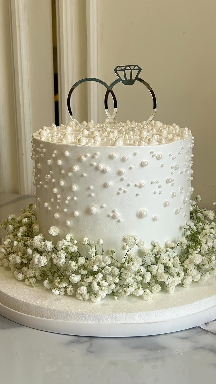 White wedding cake with pearl decorations, ring topper, and baby's breath flowers.