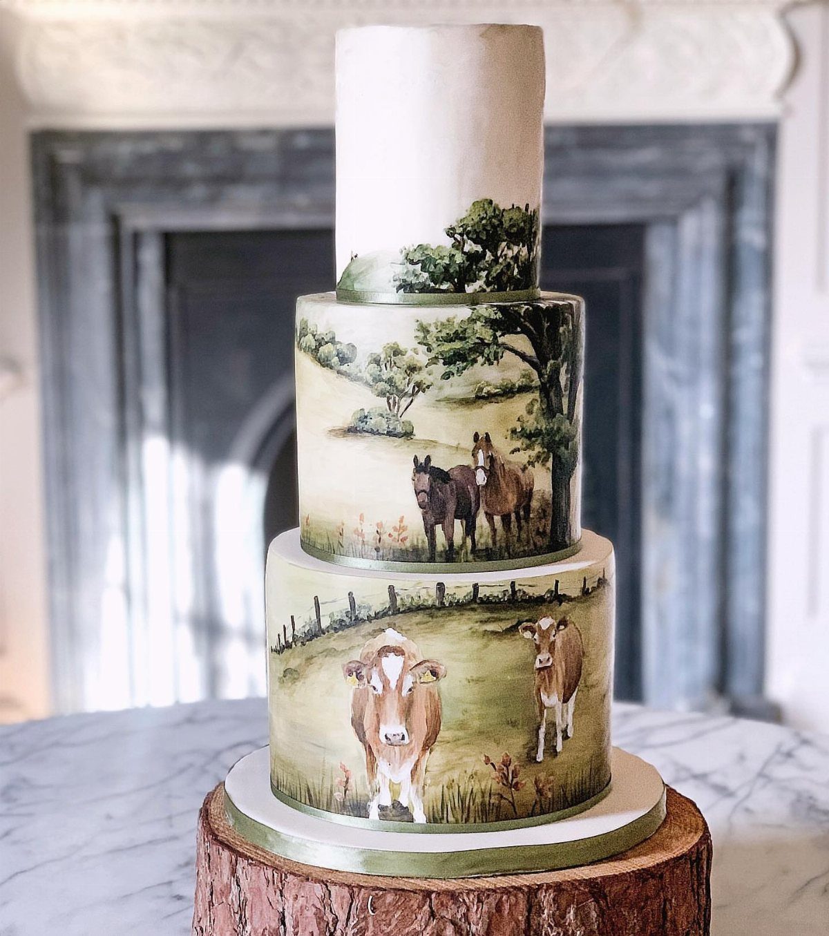 Three-tiered white cake with hand-painted farm animals and landscapes on a wooden base.