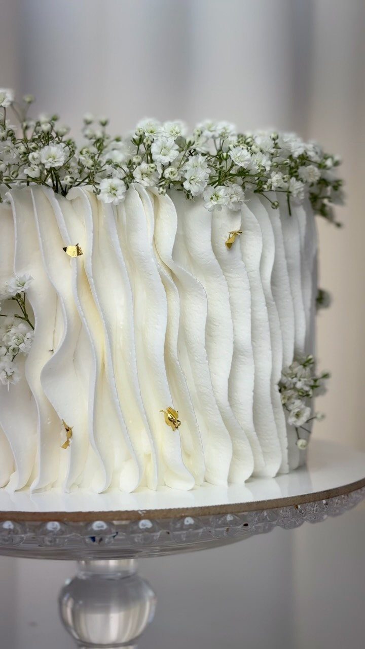 White ruffle cake topped with baby's breath flowers on a glass stand.