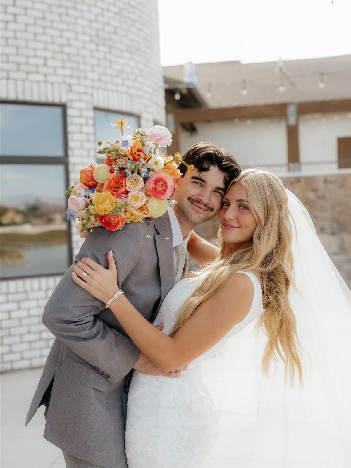 Bride hugging groom from behind with a colorful bouquet resting on his shoulder.