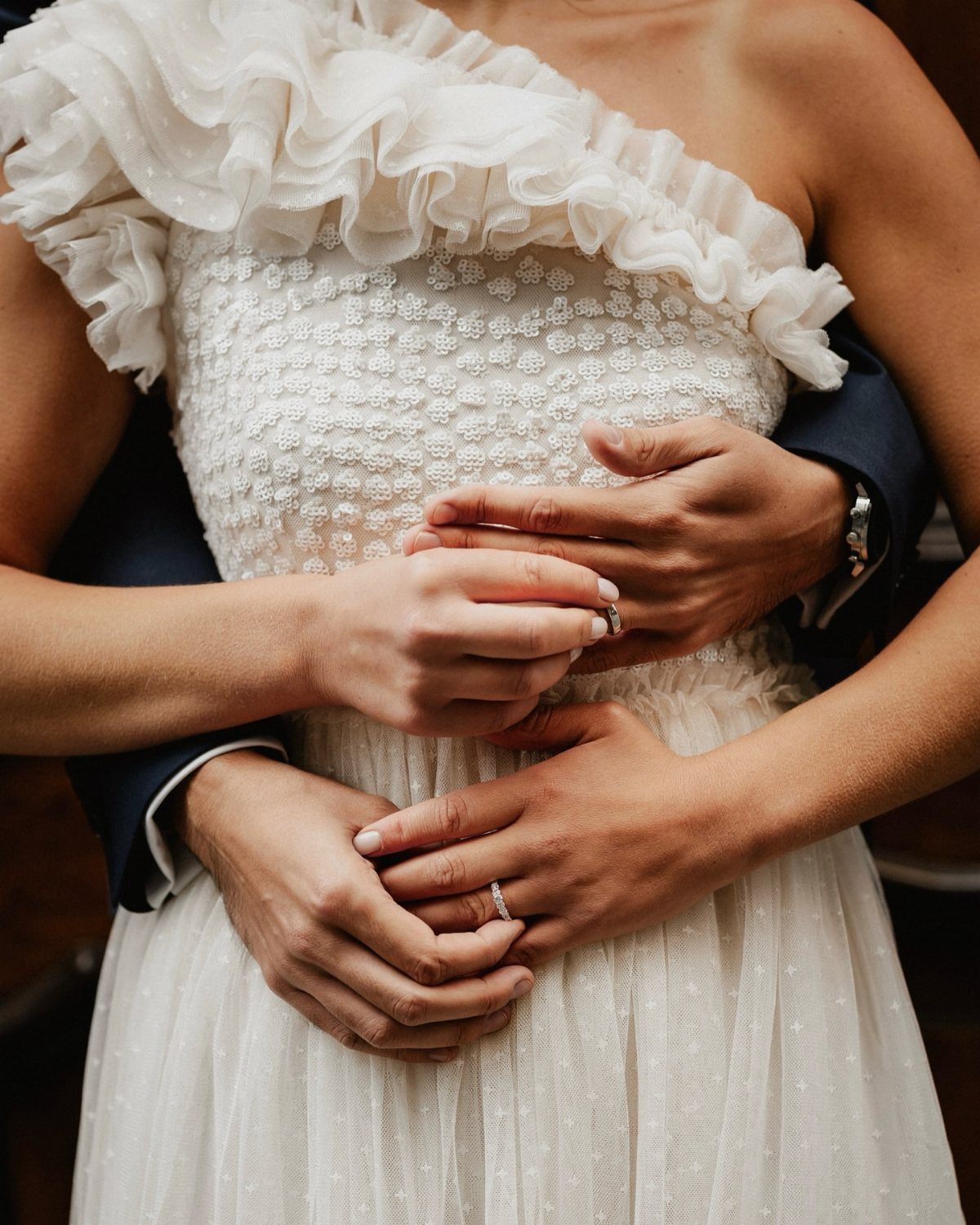 Bride in white ruffled dress embraced from behind by groom in dark suit